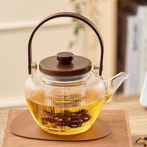 Puede incluir: Una tetera de vidrio transparente con tapa y asa de madera, llena de té. La tetera se encuentra sobre una superficie de madera y un posavasos marrón. El té es de color amarillo dorado.