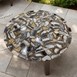 May include: A circular coffee table featuring a mosaic top composed of grey, white, and gold-toned stones. The table rests on a dark grey metal base, situated on a stone patio. The table's surface is approximately 91 cm in diameter.