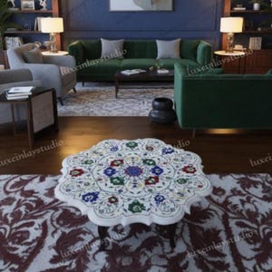 May include: A white marble coffee table with a floral inlay design in shades of red, blue, green, and yellow. The table has a scalloped edge and sits on dark wooden legs. The table is in a living room setting.
