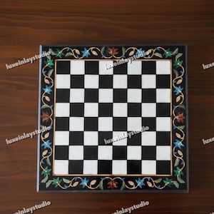 Könnte beinhalten: Quadratisches Schachbrett aus schwarzem und weißem Marmor mit floralem Rand. Der Rand hat eine goldene Umrandung und blaue, grüne und rote Blumenakzente. Das Schachbrett liegt auf einer dunklen Holzoberfläche.