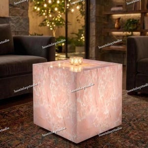 May include: A square, pink marble coffee table with a white veined pattern. The table has a soft glow and is illuminated from within. Three small candles sit on top of the table. The words "luxeinlaystudio" are printed on the side of the table.