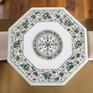 May include: An octagonal white marble table with detailed floral inlay. The table has a central circular design and a border with green and iridescent accents. The table is surrounded by a light brown rug and two chairs.