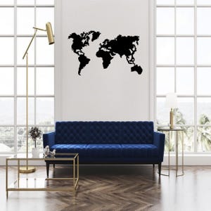 Op de afbeelding: Een zwarte wereldkaart muursticker is bevestigd op een witte muur boven een blauwe bank. De kamer heeft een modern design met een gouden vloerlamp en een salontafel met glazen blad. Grote ramen zorgen voor natuurlijk licht.