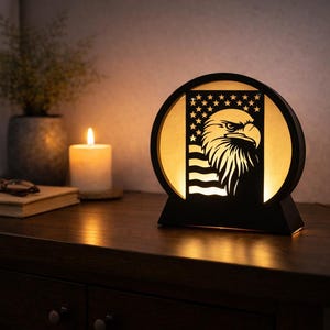 May include: A black, round, illuminated table lamp featuring a laser-cut design of an American flag and a bald eagle. The lamp is on a dark wooden surface, with a lit candle and a book in the background. The lamp is a decorative home accent.