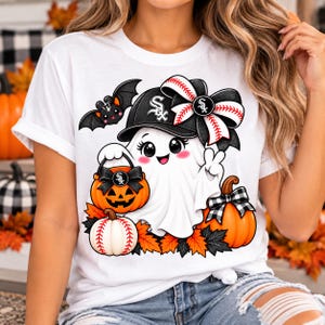 May include: A white t-shirt featuring a graphic of a cartoon ghost wearing a black baseball cap with a Chicago White Sox logo. The ghost holds an orange jack-o'-lantern bucket, accompanied by a bat, a baseball-themed bow, and pumpkins.