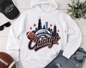 May include: A white hooded sweatshirt featuring a graphic print of the Chicago skyline, a football, and the word Chicago in navy and orange script. The design includes stylized skyscrapers, a metallic dome structure, and four orange stars.