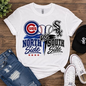 Puede incluir: Una camiseta blanca de manga corta con un diseño gráfico que incluye el logotipo de los Chicago Cubs, el logotipo de los Chicago White Sox, dos pelotas de béisbol y el texto North Side y South Side, con detalles de pinceladas azules y negras.