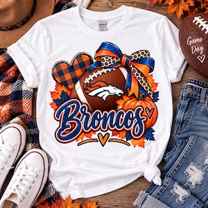 Puede incluir: Una camiseta blanca con un estampado gráfico de un balón de fútbol americano marrón con el logo de los Denver Broncos, un corazón a cuadros azul y naranja, una calabaza y un lazo grande con estampado de leopardo. El texto "Broncos" es azul.