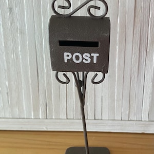 May include: A miniature brown metal mailbox with the word "POST" in white lettering. The mailbox has a rectangular body with a slot and decorative scrollwork. It stands on a thin metal pole and a square base.