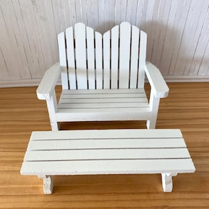 May include: A miniature white wooden bench and matching rectangular table. The bench has a slatted seat and back, with armrests. The table also has a slatted top and is supported by four legs. The furniture is designed for a dollhouse or similar setting.