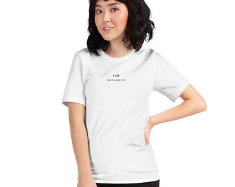 May include: A white t-shirt with the text "I AM AND SO ARE YOU" in black. The shirt is short-sleeved and worn by a person. The person is wearing black pants.