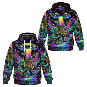 May include: A black hoodie featuring a vibrant, neon-colored graphic print of a golden Bigfoot silhouette reaching toward a glowing UFO in a lush, alien jungle landscape with purple, green, and blue foliage, palm trees, and small alien figures.