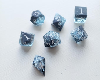 May include: A collection of seven dice for tabletop games. The dice are a mix of shapes, including a cube, a d4, a d6, a d8, a d10, a d12, and a d20. Each die has a black face with white numbers and is filled with a blue, glittery resin.
