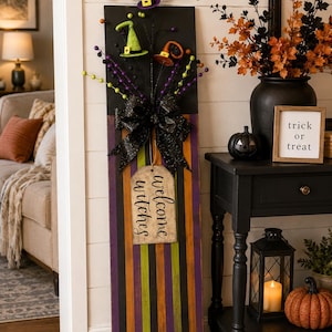 May include: A tall rectangular wooden sign featuring vertical black, purple, and green stripes. It is decorated with a large black glittery bow, three miniature witch hats in purple, green, and orange, and a hanging tag that reads welcome witches.