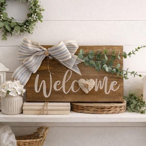 May include: A wooden sign with the word "Welcome" in white script, adorned with a striped bow and a heart. The sign is on a shelf with a white lantern, flowers, and a decorative house.