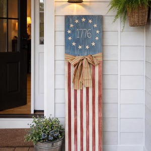May include: A wooden American flag sign with the year "1776" and stars on a blue background. The sign has red and white stripes and is decorated with a striped bow. It is displayed on a porch.
