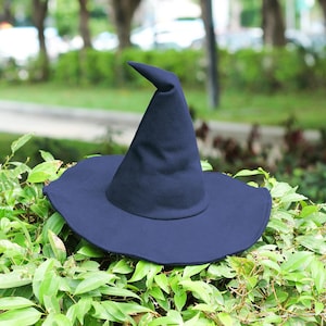 May include: A dark blue witch's hat with a broad brim and a pointed crown. The hat is made of a soft fabric and rests on green foliage, ideal for Halloween or fancy dress.