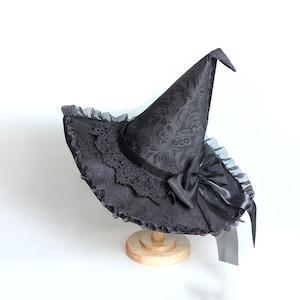 May include: A black witch's hat with a pointed crown and wide brim. The hat has a floral pattern, lace trim, and a satin bow. It is displayed on a wooden stand against a white backdrop.