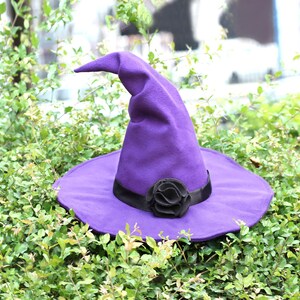 Op de afbeelding: Een paarse heksenhoed met een brede rand en een puntige kroon. De hoed heeft een zwarte lintband en een zwarte stoffen bloem. De hoed is gemaakt van een zacht materiaal en is geschikt voor Halloween of kostuumfeesten.