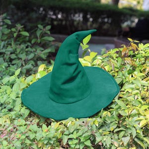 Könnte beinhalten: Ein waldgrüner Hexenhut mit spitzer Spitze und breiter, flatternder Krempe. Der Hut ist aus weichem Stoff gefertigt und vor dem Hintergrund grüner Blätter platziert. Ideal für Halloween oder Kostümpartys.