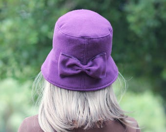 Puede incluir: Un sombrero cloche morado con un lazo decorativo en la parte trasera. El sombrero tiene una copa redonda y un ala. El fondo es un verde borroso, lo que sugiere un entorno al aire libre. El sombrero está hecho de un material suave.