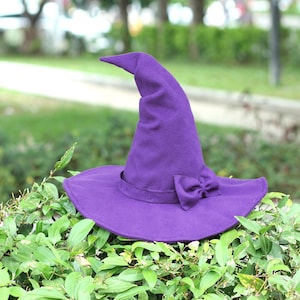 Op de afbeelding: Een felpaarse heksenhoed met een brede rand en een decoratieve strik. De hoed is gemaakt van een zachte stof en staat tegen een achtergrond van groen gebladerte. Een klassiek Halloween-kostuumaccessoire.