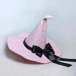 May include: A light pink pointed hat with a subtle embossed floral pattern throughout the fabric. The hat features a wide, slightly wavy brim and a tall, conical crown. A smooth, black satin ribbon is tied around the base into a bow.