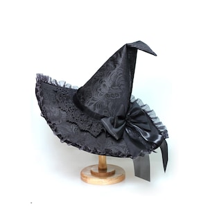 Peut inclure: Un chapeau de sorcière noir avec une couronne pointue et un large bord. Le chapeau présente un motif floral, une bordure en dentelle et un grand nœud en satin. Il est présenté sur un support en bois sur fond blanc.