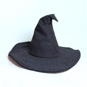 May include: A dark grey witch's hat with a wide brim and a pointed crown. The hat is made of a soft, felt-like material, suitable for Halloween or costume parties. The hat is set against a white background.