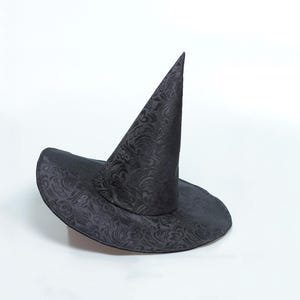May include: A black pointed hat featuring a tonal, intricate floral damask pattern throughout the fabric. The hat has a wide, circular brim and a tall, conical crown that tapers to a sharp point, creating a structured silhouette.