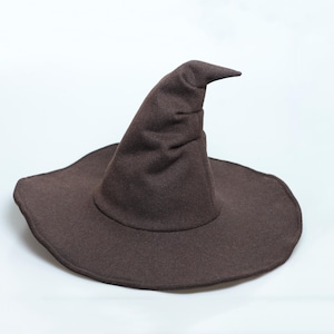 May include: A dark brown witch hat featuring a wide, circular brim and a tall, pointed crown that curves slightly at the top. The hat is constructed from a soft, matte fabric with visible stitching along the edge of the brim and the base of the crown.