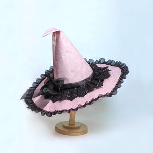 以下が含まれることがあります： 淡いピンク色の魔女の帽子。同系色の繊細な花柄が施され、つばには黒いフリル、帽子の付け根には黒いレースのオーバーレイと黒いリボンの装飾があしらわれています。帽子は小さな木製のスタンドに飾られています。