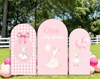May include: Custom stretchy arch covers/stands with a pink theme. The arches feature illustrations of geese, bows, and balloons. One arch has the text "One silly goose". The arches are set up on a grassy lawn with pink balloons.