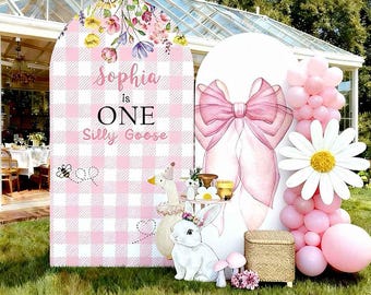 May include: A pink and white checkered arch cover with the text "Sophia is ONE Silly Goose". The arch is decorated with floral arrangements, a large pink bow, and cartoon animal figures. A daisy and balloons are also present.