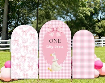 May include: Three pink arch-shaped party decorations. The center arch features a pink bow, the words "ONE Silly Goose", and a cartoon goose. The other arches have pink bow and gingham patterns. The text "Custom Stretchy Arch Cover/stand" is at the top.