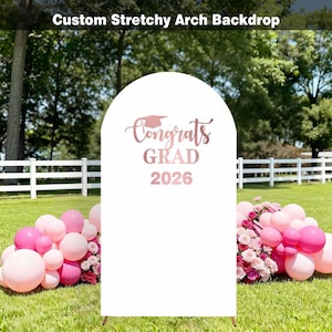 May include: A white, arched backdrop with the text "Congrats GRAD 2026" in rose gold. The backdrop is set on a grassy lawn, decorated with pink balloons and flowers. The top of the backdrop reads "Custom Stretchy Arch Backdrop".