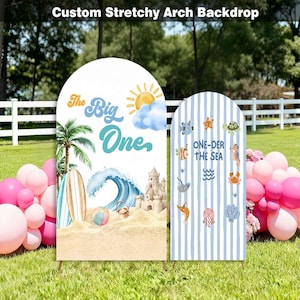 May include: Two arched fabric backdrops featuring beach-themed graphics. The left backdrop displays a palm tree, surfboard, ocean wave, sandcastle, and the text 'The Big One'. The right backdrop has blue stripes with various sea creature illustrations.