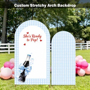 May include: A custom stretchy arch backdrop with a blue and white gingham pattern. One arch features the text "She's Ready to Pop!" with a soda bottle illustration and a blue bow. Pink and white balloons are in the foreground.