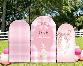 May include: Pink arch covers with a gingham pattern and bow accents. One cover features the text "ONE Silly Goose" with a cartoon goose illustration. The covers are set up on a grassy lawn with pink balloons.