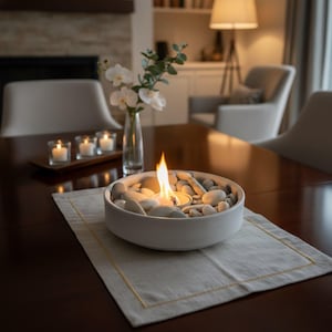May include: A white ceramic bowl filled with smooth, light-colored stones, with a small flame burning in the center. The bowl sits on a beige placemat on a dark wooden table. Other decor includes candles and a vase of flowers.