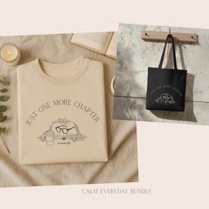 Puede incluir: Una sudadera beige y un bolso tote negro con el texto "JUST ONE MORE CHAPTER" y un diseño de libro. La sudadera tiene el mismo diseño. Una vela encendida, una ramita de vegetación y un libro abierto también están en la imagen.
