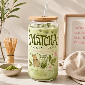Może przedstawiać: Przezroczysta szklanka wypełniona mrożoną matchą latte, z bambusową pokrywką i szklaną słomką. Na zewnątrz znajduje się zielony napis "MATCHA SOCIAL CLUB" oraz "A little matcha, a lot of magic" wraz z ilustracjami pędzelka i liści herbaty.