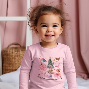 May include: A light pink long-sleeve shirt featuring a graphic print of a Christmas tree, a nutcracker, two ballerinas, gifts, and candy canes. The text "Let the christmas magic begin" is printed in a cursive font at the bottom of the graphic.