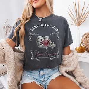 Könnte beinhalten: Dunkelgraues T-Shirt mit dem Schriftzug "DARK ROMANCE" in einem Bogen. Darunter ein offenes Buch mit Rosen, einem Totenkopf und dem Schriftzug "Readers Society". Der Text "Midnight Chapters & Morally Grey Hearts" ist ebenfalls vorhanden.