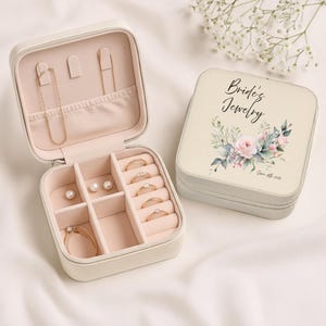 Könnte beinhalten: Eine cremefarbene Schmuckschatulle, geöffnet, um einen rosa Innenraum mit Fächern für Ringe, Halsketten und Ohrringe zu zeigen. Daneben eine geschlossene Schatulle mit dem Text "Bride's Jewelry" und floralem Design. Der Schmuck umfasst Ringe, eine Halskette und Ohrringe.