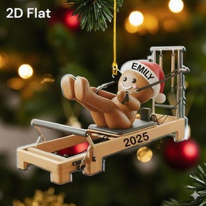 Op de afbeelding: Een 2D platte kerstversiering met een peperkoeken mannetje met een kerstmuts met de naam "Emily" en het jaar "2025" op een Pilates reformer. De versiering is bruin, beige en rood, met een gouden ophangkoord.