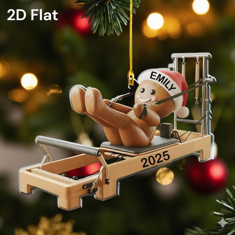 Op de afbeelding: Een 2D platte kerstversiering met een peperkoeken mannetje met een kerstmuts met de naam "Emily" en het jaar "2025" op een Pilates reformer. De versiering is bruin, beige en rood, met een gouden ophangkoord.