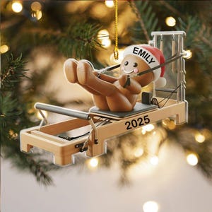 Op de afbeelding: Kerstornament met een peperkoeken mannetje met een kerstmuts, die oefent op een Pilates reformer. Het ornament is bruin met zwarte en witte accenten. Het jaar "2025" staat op de machine.