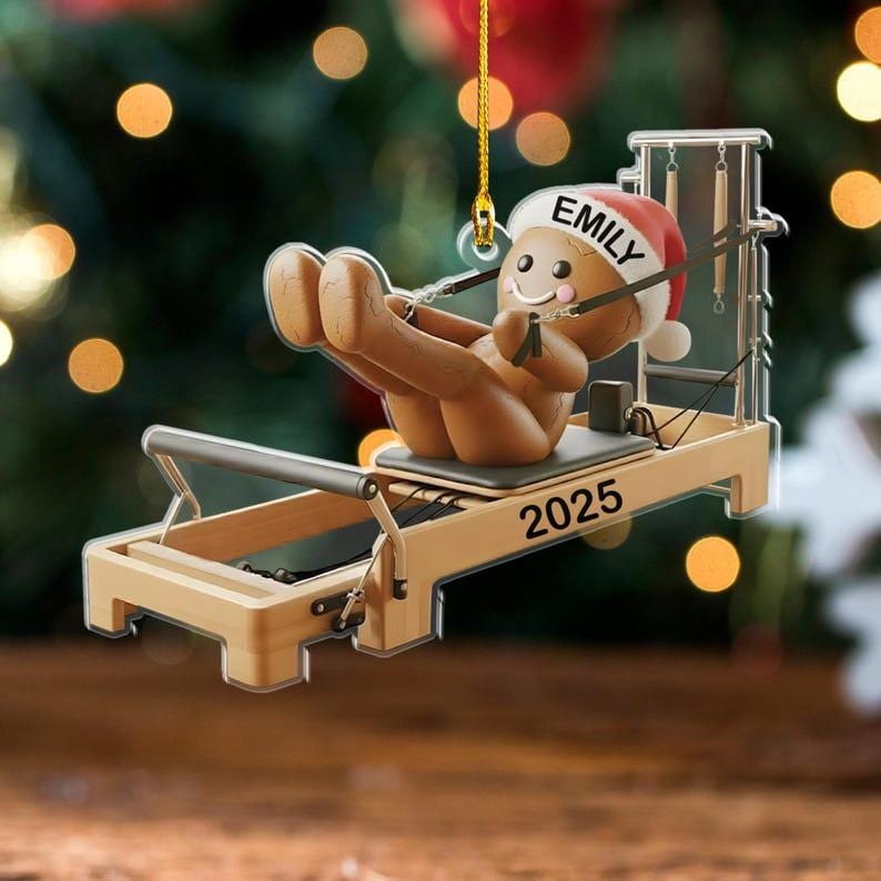 Op de afbeelding: Kerstboomversiering met een peperkoeken man die Pilates doet op een apparaat. De versiering heeft een kerstmuts met de naam "EMILY" en het jaar "2025". Opgehangen aan een gouden draad.