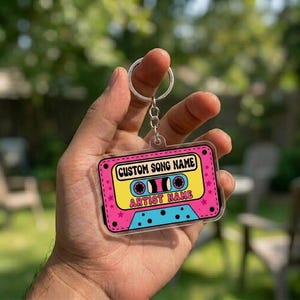 May include: A retro-style keychain featuring a cassette tape design. The keychain has a pink border with yellow and blue details. Text on the keychain reads "CUSTOM SONG NAME" and "ARTIST NAME". A silver key ring is attached.